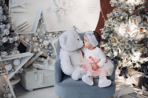 Schattig babymeisje met teddybeer op fauteuil bij kerstboom.