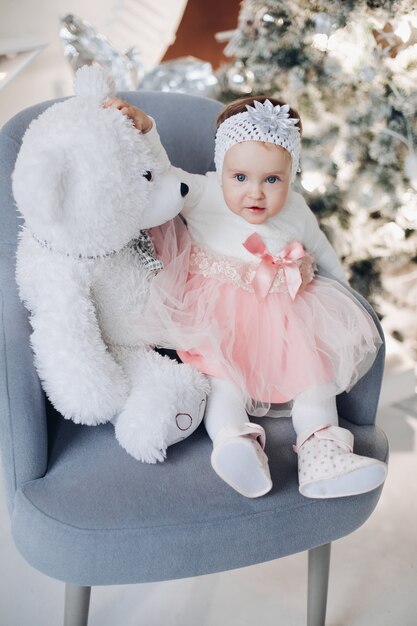 Schattig babymeisje met teddybeer op fauteuil bij kerstboom.