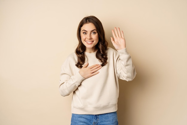 Schattig bescheiden meisje glimlachend opstekende vinger stelt zichzelf voor en zegt hallo terwijl ze over een beige achtergrond staat