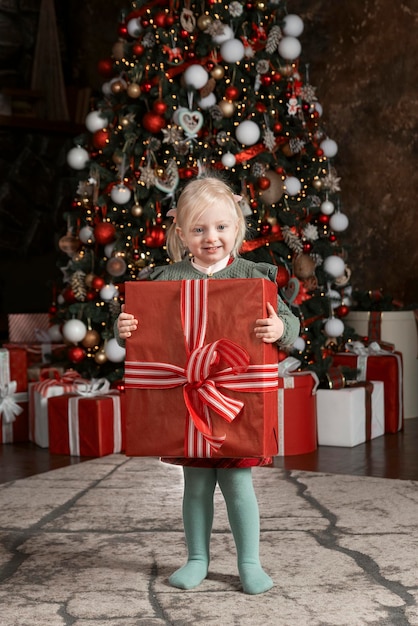 Schattig blond kindmeisje met een enorme rode geschenkdoos Klein meisje met een groot cadeau op de achtergrond van de kerstboom