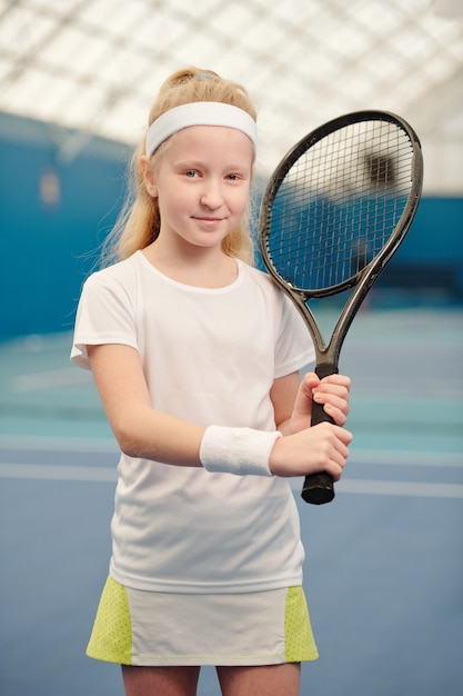 Schattig blond meisje in activewear die tennisracket tegen de linkerschouder houdt terwijl ze voor de camera in de stadionomgeving staat