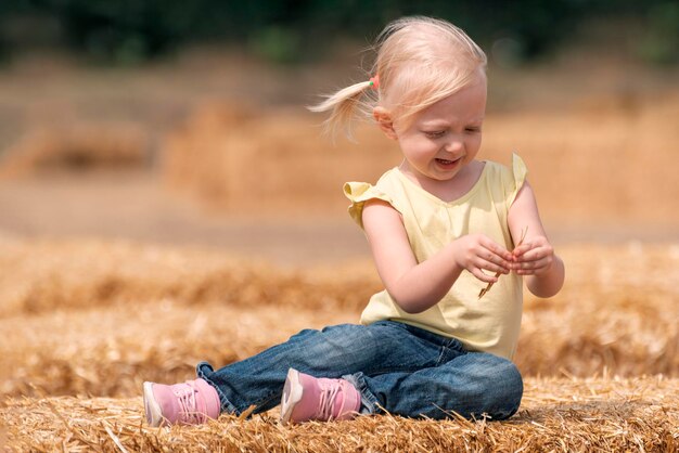 Schattig blond meisje met paardenstaarten zit in het hooi. Weekendje op de boerderij. Portret van een kind op hooiberg.