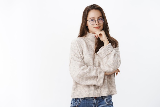 Schattig brunette meisje met een bril en een gezellige trui die de hand op de kin houdt en glimlacht