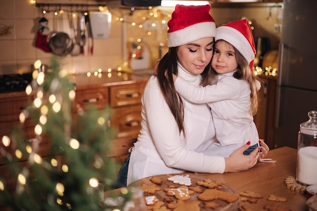 Schattig dochtertje met haar moeder in de keuken tijdens de kerstvakantie, decoratieve geschikte boom bij