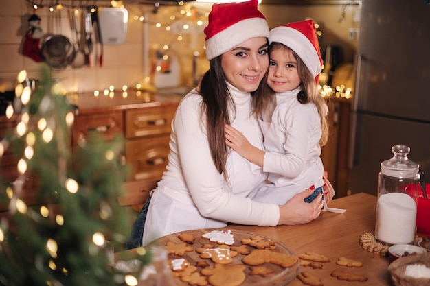 Schattig dochtertje met haar moeder in de keuken tijdens de kerstvakantie, decoratieve geschikte boom bij