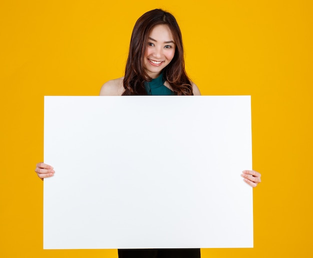 Schattig en mooi krullend haar aziatische vrouwelijke brunette houden en wijzend op wit leeg bord poseert voor camera met een vrolijke voor reclame en banner gebruiksdoel, geïsoleerd op felgele achtergrond