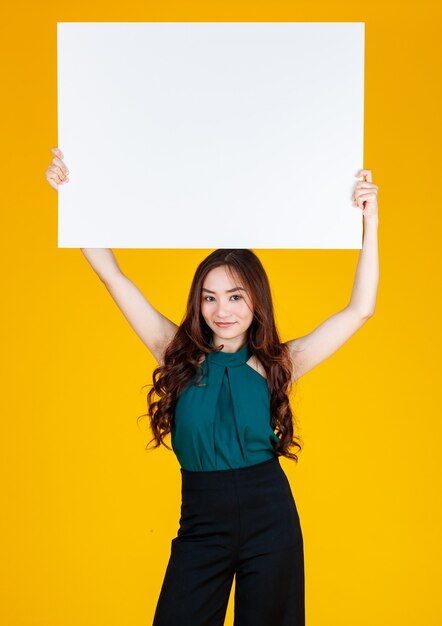 Schattig en mooi krullend haar Aziatische vrouwelijke brunette met een wit leeg bord boven haar hoofd met een vreugdevol voor reclame- en bannergebruiksdoel, studio-opname geïsoleerd op felgele achtergrond.