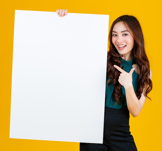 Schattig en mooi krullend haar Aziatische vrouwelijke brunette met wit leeg bord poseert voor de camera met een vreugdevol voor reclame- en bannergebruiksdoel, studio-opname geïsoleerd op felgele achtergrond.