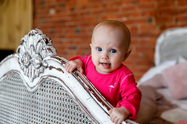 Schattig gelukkig blauwogig 6 maanden oud meisje in felrode jurk aan het hoofd van retro bed. Vrij lachende blonde schoonheid. Charmant kind toont emoties in close-up. Concept van gezond kind en ouderschap