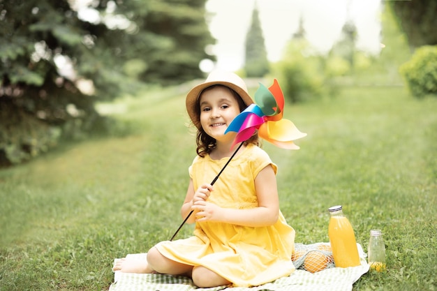 Schattig gelukkig klein meisje in een hoed met plezier in parkweide Kind zittend op een deken met pinwheel en kijken naar camera picknick op de natuur