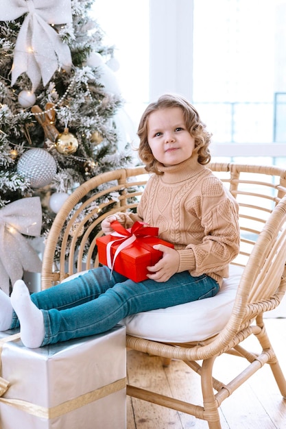 schattig gelukkig klein meisje met krullen zit op een stoel in de buurt van de kerstboom met geschenkdoos in haar handen