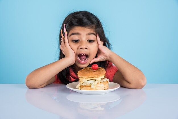 Schattig Indiaas of Aziatisch meisje dat smakelijke hamburger, sandwich of pizza eet in een bord of doos. Staande geïsoleerd over blauwe of gele achtergrond.