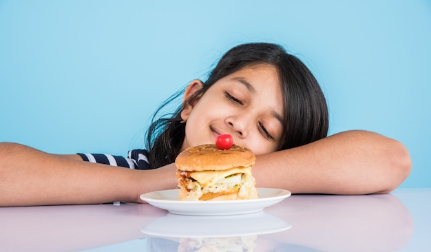 Schattig Indiaas of Aziatisch meisje dat smakelijke hamburger, sandwich of pizza eet in een bord of doos. Staande geïsoleerd over blauwe of gele achtergrond.