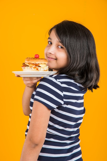 Schattig Indiaas of Aziatisch meisje dat smakelijke hamburger, sandwich of pizza eet in een bord of doos. Staande geïsoleerd over blauwe of gele achtergrond.