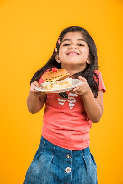 Schattig Indiaas of Aziatisch meisje dat smakelijke hamburger, sandwich of pizza eet in een bord of doos. Staande geïsoleerd over blauwe of gele achtergrond.