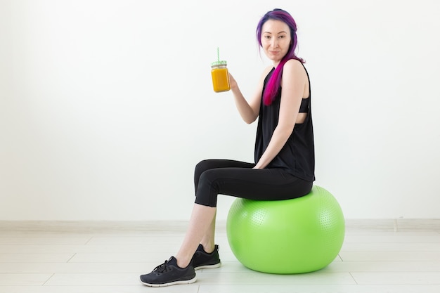 Schattig jong gemengd ras hipster meisje met gekleurd haar zittend op een groene fitball en met een banaan eiwit smoothie in haar handen op een witte achtergrond. Gezond eten en bewegen concept. Copyspace