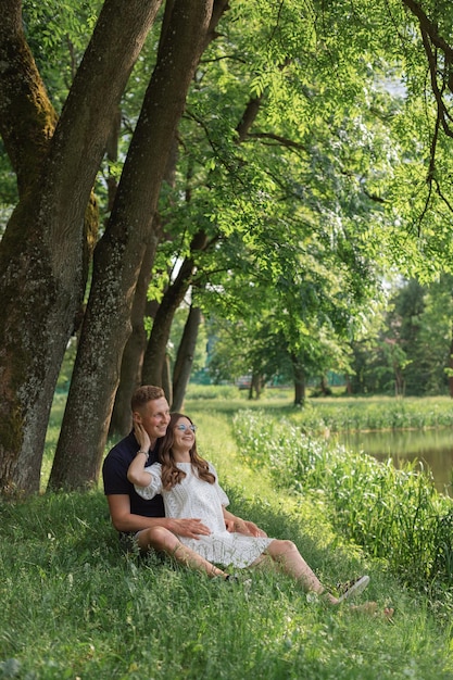 Schattig jong koppel zittend op het gras onder de boom in de buurt van het meer kijkend naar elkaar jonge man vrouw enjoyi
