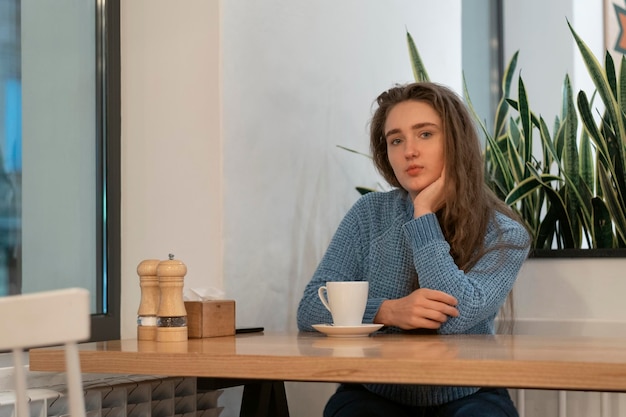 Schattig jong meisje met bruin haar in blauwe gebreide trui zit in café met kopje thee en kijkt bedachtzaam in de camera