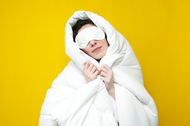 Schattig jong meisje slaapt in slaapmasker en bedekt zichzelf met een zachte warme deken op gele achtergrond