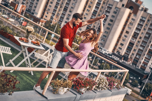 Schattig jong stel in casual kleding dansen en glimlachen terwijl ze een romantisch diner hebben op het dakterras buiten