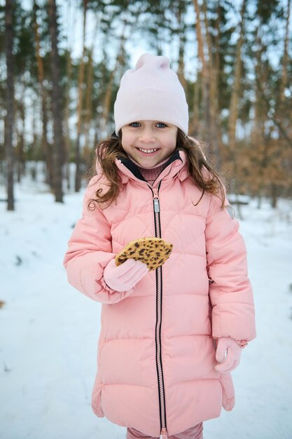 Schattig kind mooi meisje in roze pastel warme winterkleren met heerlijke koekjes met chocolade snacken buiten in een besneeuwde natuur Geniet van wintervakanties en ontdek de natuur