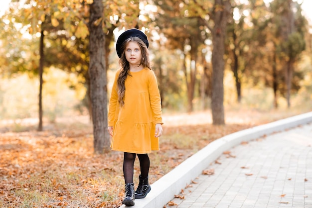 Schattig kindmeisje van 5-6 jaar oud draagt een gele jurk en een baret die op straat in de stad loopt over de herfstnatuur