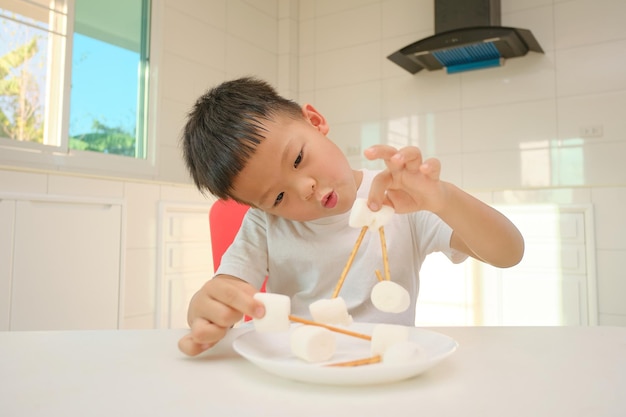 Foto schattig klein aziatisch jongenskind dat aan de keukentoren zit met thuisonderwijs met marshmallows