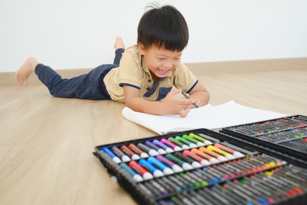 Schattig klein Aziatisch kleuterjongenskind dat op zijn buik op de vloer ligt terwijl hij tekent met kleurrijke kleurpotloden en veel tekengereedschappen thuis Creatief spel voor kind verbetert de focus in het kinderconcept