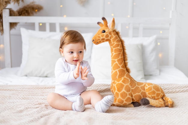 Schattig klein babymeisje met een gevuld girafspeelgoed zit op een bed met een lichte kamer na het slapen en glimlachend in haar handen klappend