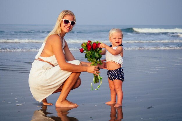 Foto schattig klein blond meisje dat moeder een boeket rozen geeft 's ochtends op het strand bij de zee. moederdag en verjaardagsviering. valentijnsdag 14 februari mooie vrouw en haar dochter oceaanvakantie