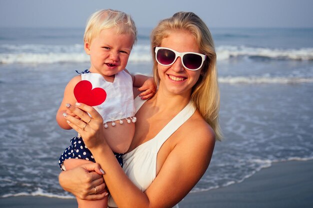 Schattig klein blond meisje en moeder met kleine papieren harten 's ochtends op het strand bij de zee. Moederdag en verjaardagsviering. Valentijnsdag 14 februari mooie vrouw en dochter oceaanvakantie