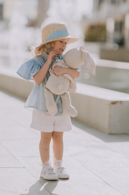 schattig klein blond meisje in blauwe jurk en strohoed spelen in de buurt van fontein met pluche konijntje, kaart