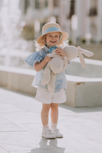 Schattig klein blond meisje in blauwe jurk en strohoed spelen in de buurt van fontein met pluche konijntje, kaart