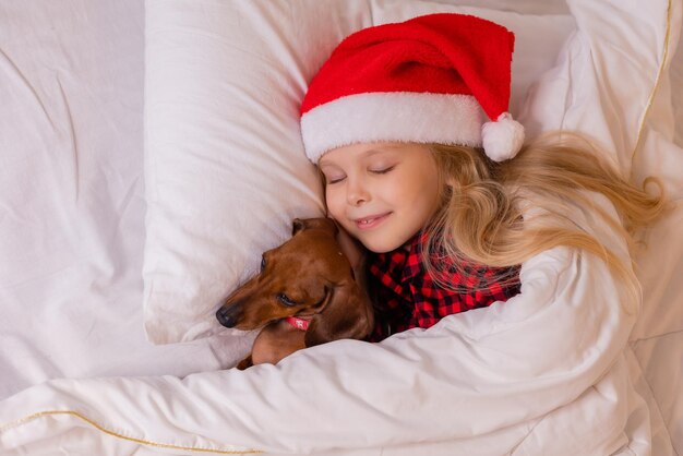 schattig klein blond meisje in een kerstmuts en pyjama slaapt in bed met haar geliefde hond