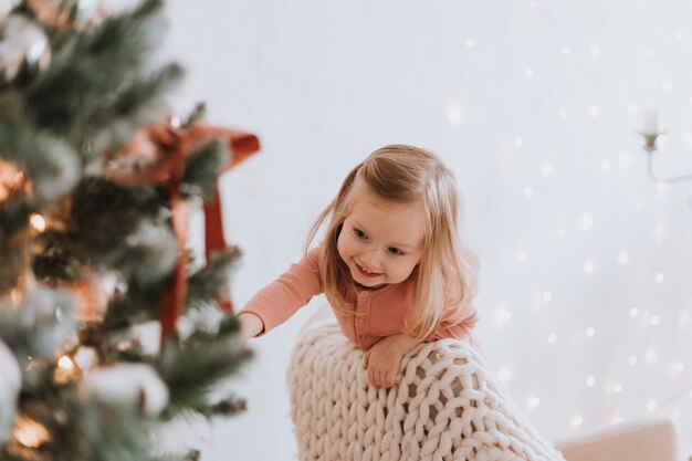 schattig klein blond meisje knielt in een stoel tegen de achtergrond van een kerstboom