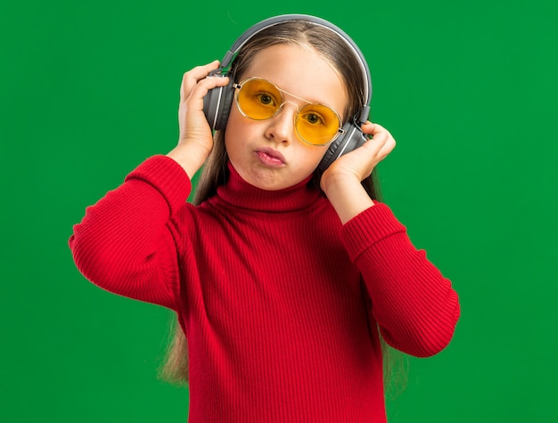 Schattig klein blond meisje met een koptelefoon en een zonnebril die een koptelefoon grijpt en naar een camera kijkt die op een groene muur is geïsoleerd