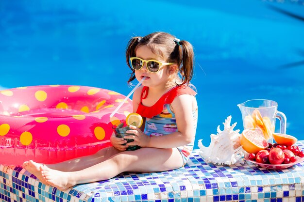 schattig klein brunette meisje in zonnebril en zwembroek drinkt limonade bij het zwembad.