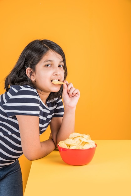 Schattig klein indiaans of Aziatisch meisje dat chips of aardappelwafels eet in grote rode kom, over gele achtergrond