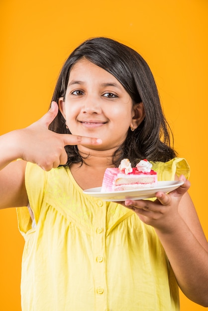 Schattig klein Indiaas of Aziatisch meisje dat een stuk aardbei of chocoladegebak of cake in een bord eet. Geïsoleerd over kleurrijke achtergrond