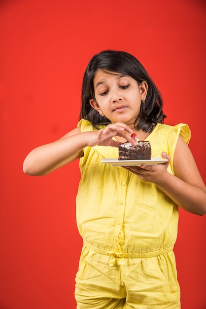 Schattig klein indiaas of aziatisch meisje dat een stuk aardbei of chocoladegebak of cake in een bord eet. geïsoleerd over kleurrijke achtergrond