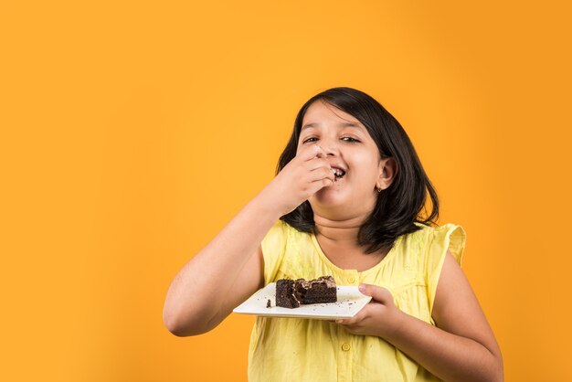 Schattig klein Indiaas of Aziatisch meisje dat een stuk aardbei of chocoladegebak of cake in een bord eet. Geïsoleerd over kleurrijke achtergrond