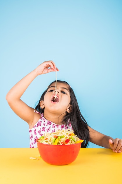 Schattig klein Indiaas of Aziatisch meisje dat lekkere Chinese noedels eet met vork of eetstokjes, geïsoleerd over kleurrijke achtergrond colorful