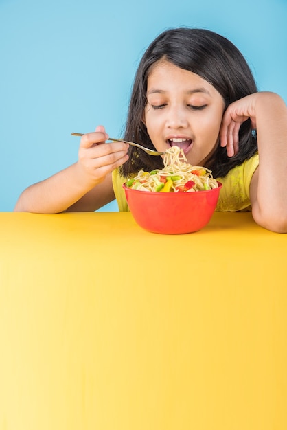 Schattig klein Indiaas of Aziatisch meisje dat lekkere Chinese noedels eet met vork of eetstokjes, geïsoleerd over kleurrijke achtergrond colorful