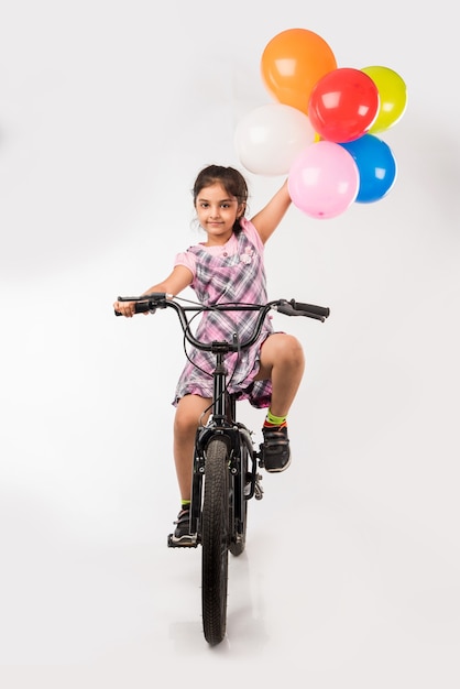 Schattig klein Indiaas of Aziatisch meisje dat op de fiets rijdt, geïsoleerd op een witte achtergrond met ballonnen