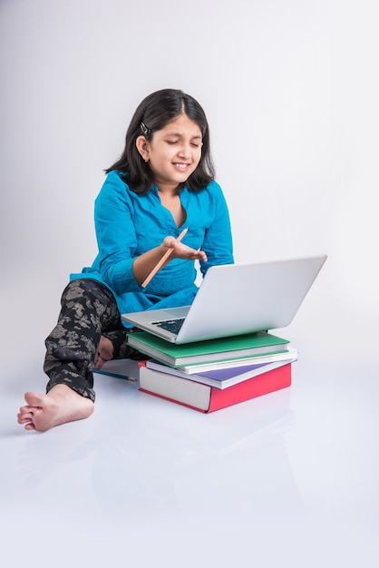 Schattig klein Indiaas of Aziatisch meisje dat op laptop studeert of aan een schoolproject werkt terwijl liggend of zittend op de vloer, geïsoleerd op witte achtergrond