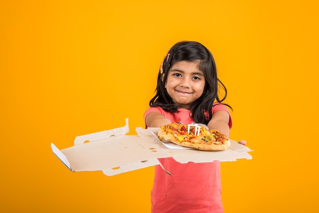 Schattig klein Indiaas of Aziatisch meisje dat smakelijke hamburger, sandwich of pizza eet in een bord of doos. Staande geïsoleerd over blauwe of gele achtergrond.