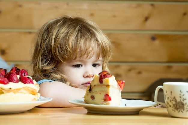 Schattig klein jongenskind met lang blond haar dat smakelijke romige taart of cake eet met rood aardbeienfruit...