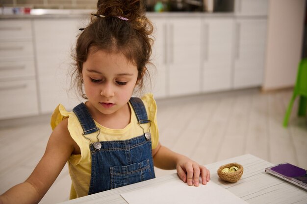 Schattig klein Kaukasisch meisje in geel t-shirt en blauw denim dat thuis aan tafel zit en foto's maakt