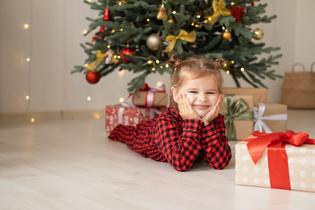 schattig klein kind meisje in rode pyjama liggend op de vloer in de buurt van kerstboom met geschenkdozen thuis