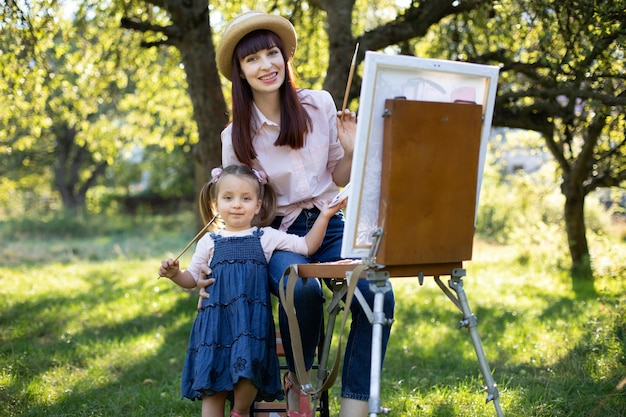 Schattig klein kindmeisje in de zomertuin, houdt een penseel vast en schildert samen met haar moeder een afbeelding op canvas. Mooie jonge moeder zit op de stoel en knuffelt het meisje